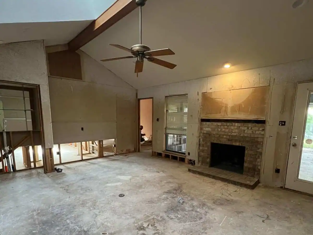 efore—Dallas living room mid-remodel showing brick fireplace, exposed framing, subfloor, and ceiling fan